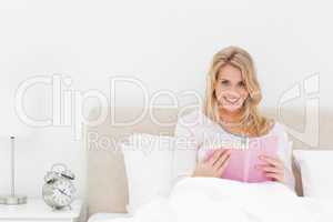 Woman in bed reading a book, while looking forward and smiling