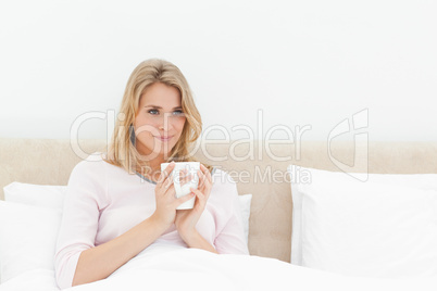 Woman in bed with a cup in hands, softly smiling while looking t