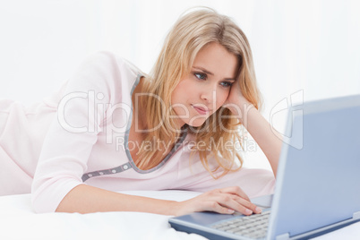 Woman lying on bed scrolling on her laptop while resting her hea