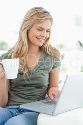 Woman smiling, as she uses her laptop and hold a cup in her othe