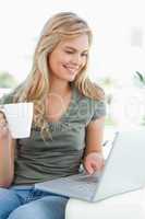 Woman smiling, as she uses her laptop and hold a cup in her othe