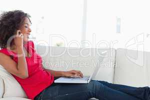 Woman making a call and using her laptop while on the couch smil