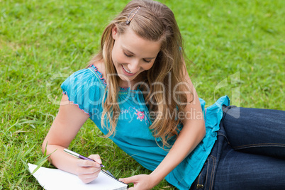 Young smiling girl lying on the grass while writing on a noteboo