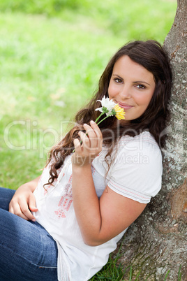 Young smiling woman smelling flowers while sitting against a tre