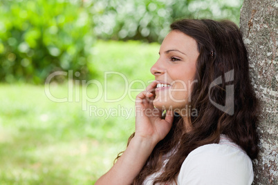 Young smiling woman leaning against a tree while talking on the