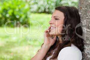 Young smiling woman leaning against a tree while talking on the