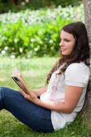 Young thoughtful girl leaning against a tree while reading a boo