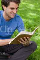 Young smiling man reading a book while siting on the grass