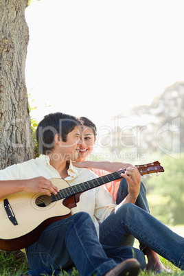 Man playing a guitar and sitting against a tree while his friend