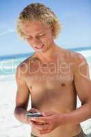 Smiling blonde man standing on the beach while sending a text
