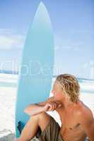 Serious blonde man sitting on the beach with his surfboard next