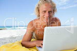 Smiling blonde man lying on the beach while using his laptop