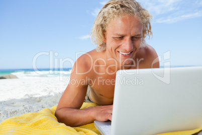 Smiling blonde man looking at his laptop while lying on the beac