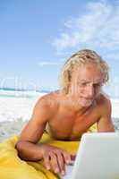 Blonde man lying on the beach while using his laptop in a very c