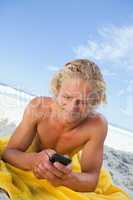 Blonde man lying on his beach towel while sending a text
