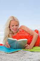 Young smiling woman lying on her beach towel while reading a boo