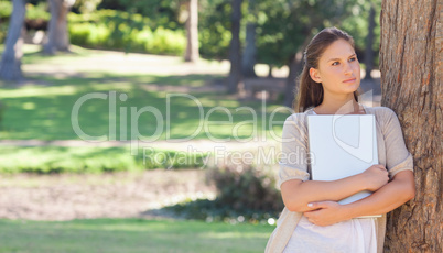 Woman holding a laptop while leaning against a tree