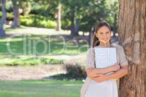 Smiling woman with a laptop leaning against a tree