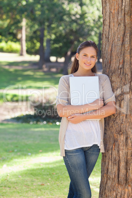 Smiling woman holding a laptop while leaning against a tree