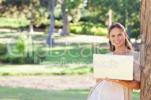 Smiling woman using a notebook while leaning against a tree