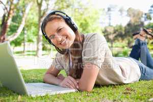 Smiling woman with a headset and a laptop lying on the lawn