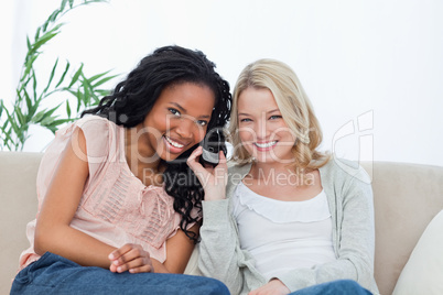 Two women listening to a mobile phone are smiling at the camera