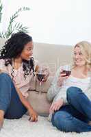 Two women sitting up against a couch are drinking wine