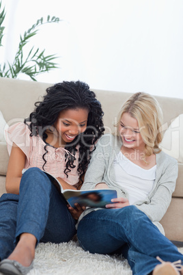 Two smiling women are sitting on the ground reading out of a mag