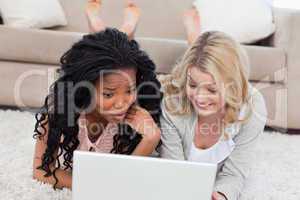 Two women using a laptop are lying on the ground