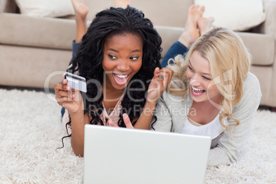 Two laughing women are lying on the floor with a laptop and a ba