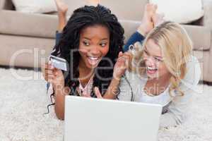 Two laughing women are lying on the floor with a laptop and a ba
