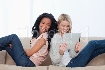 Two women sitting back to back on a couch looking at a tablet co