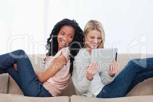 Two women sitting back to back on a couch looking at a tablet co