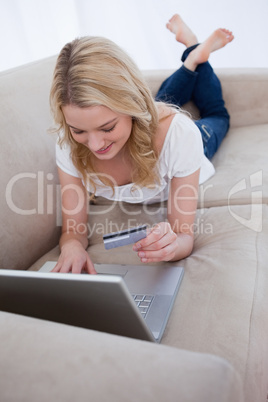 A woman lying on a couch is holding her bank card and typing on