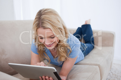 A smiling woman lying on a couch is using her tablet