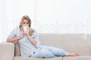 A woman drinking a cup of coffee is lying on a couch
