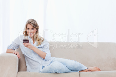 A smiling woman lying on a couch is holding a glass of wine