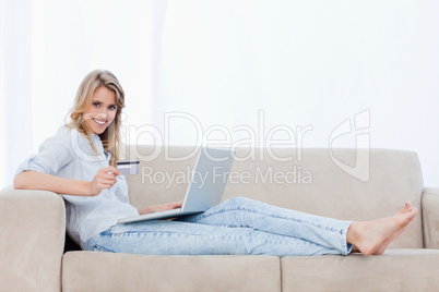 A woman holding a bank card is lying on a couch with a laptop in