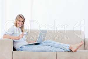 A woman holding a bank card is lying on a couch with a laptop in
