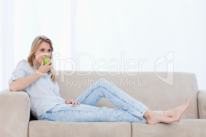 A woman lying on a couch is eating an apple