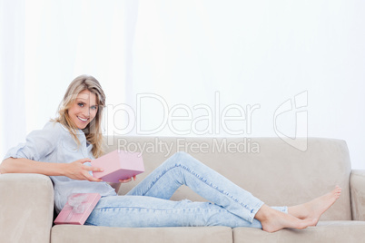A smiling woman lying on a couch is holding a pink box