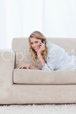 A smiling woman talking on her mobile phone is lying on a couch