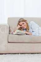 A woman looking at the camera is lying on a couch with a book