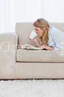 A woman lying on a couch with a book is drinking coffee