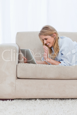 A woman lying on a couch holding a tablet is laughing