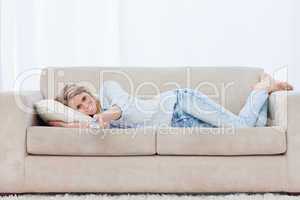 A smiling woman lying on a couch is holding a TV remote control