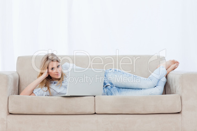 A woman looking at the camera is lying on a couch using her lapt