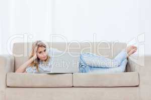 A woman looking at the camera is lying on a couch using her lapt