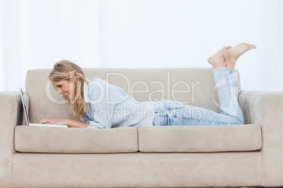 Side view shot of a smiling woman lying on a couch typing on her