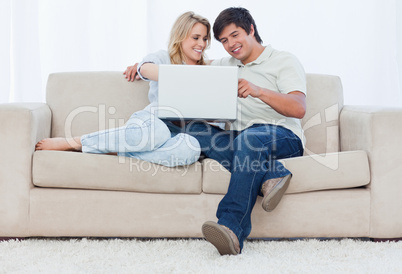 A smiling couple are sitting on a couch looking at a laptop